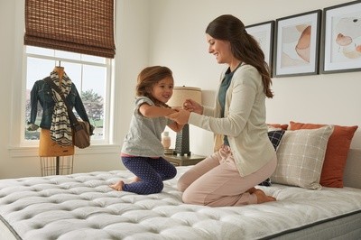 woman kneeling on bed holding little girl's hands while she bounces on the bed